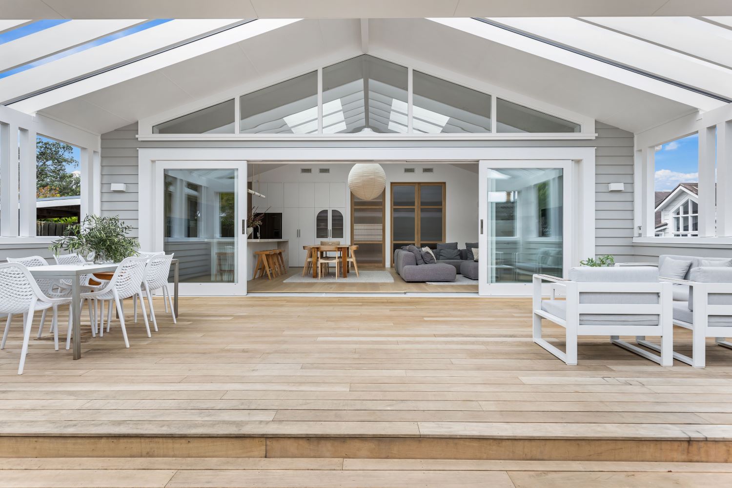 Modern outdoor patio with wooden deck, white outdoor furniture, and dining set, leading into a bright, open-plan living and kitchen area through large sliding glass doors.